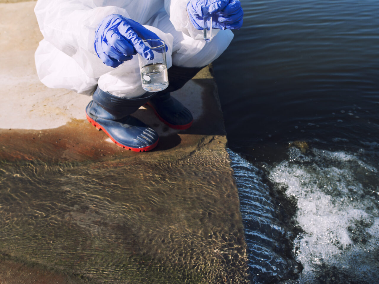 Unrecognizable ecologist standing where sewage waste water meets the river and taking samples to determine level of contamination and pollution.