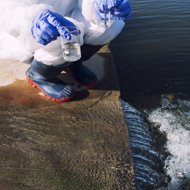 Unrecognizable ecologist standing where sewage waste water meets the river and taking samples to determine level of contamination and pollution.